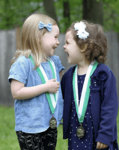 Two Delphi Academy students share a joyful laugh while wearing academic achievement medals, demonstrating the positive outcomes of goal setting for kids. The girls' genuine excitement and matching green medals represent successful goal completion and peer celebration, key aspects of Delphi's goal-setting approach.